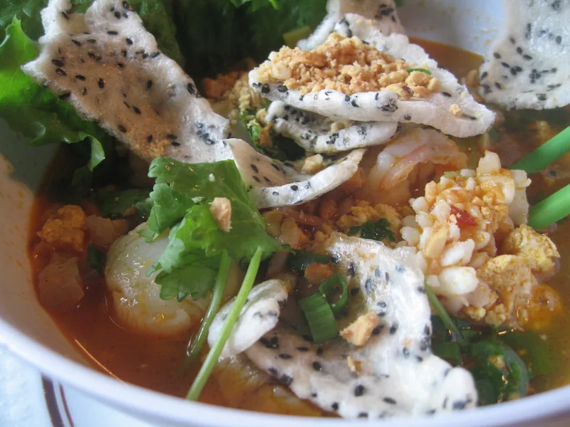 A bowl of Da Nang's mi quang noodle soup topped with shrimp, herbs, peanuts, and sesame rice crackers.