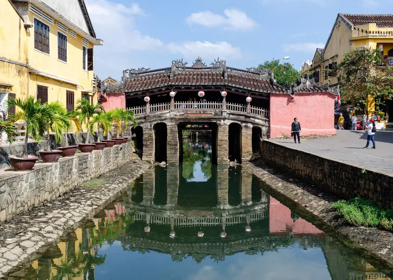 The historic covered Japanese Bridge in Hoi An reflects in the canal under a sunny sky.
