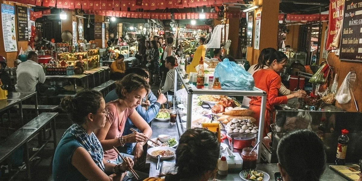Diners sit at a busy food stall eating noodles under lanterns in a Hoi An market.