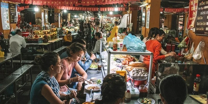 Diners sit at a busy food stall eating noodles under lanterns in a Hoi An market.