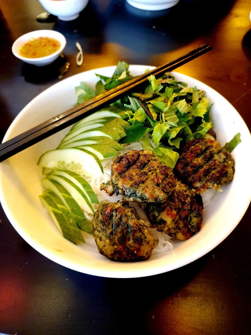 A bowl of Vietnamese bun cha with grilled pork patties, rice noodles, cucumber and herbs, served with dipping sauce.