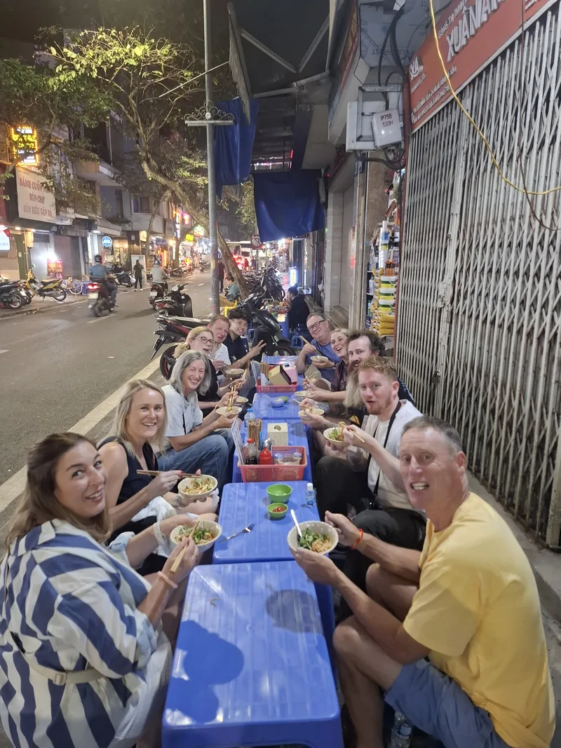 Tourists sit on plastic stools sharing bowls of noodles at a street food stall in Hanoi's Old Quarter at night.