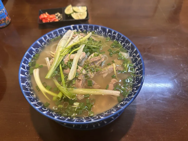 A bowl of Vietnamese beef pho noodle soup topped with scallions and herbs, served with lime and chili on the side.