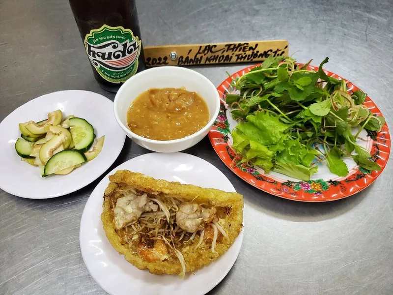 A Hue table spread of crispy banh khoai, fresh herbs, pickled vegetables, peanut sauce and a bottle of Huda beer.