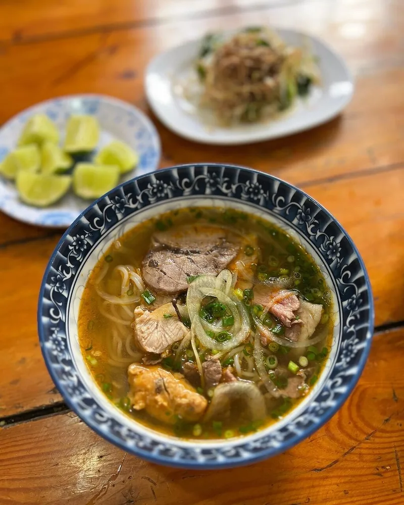 A bowl of Hue beef noodle soup with sliced meat and herbs served alongside lime wedges on a wooden table.