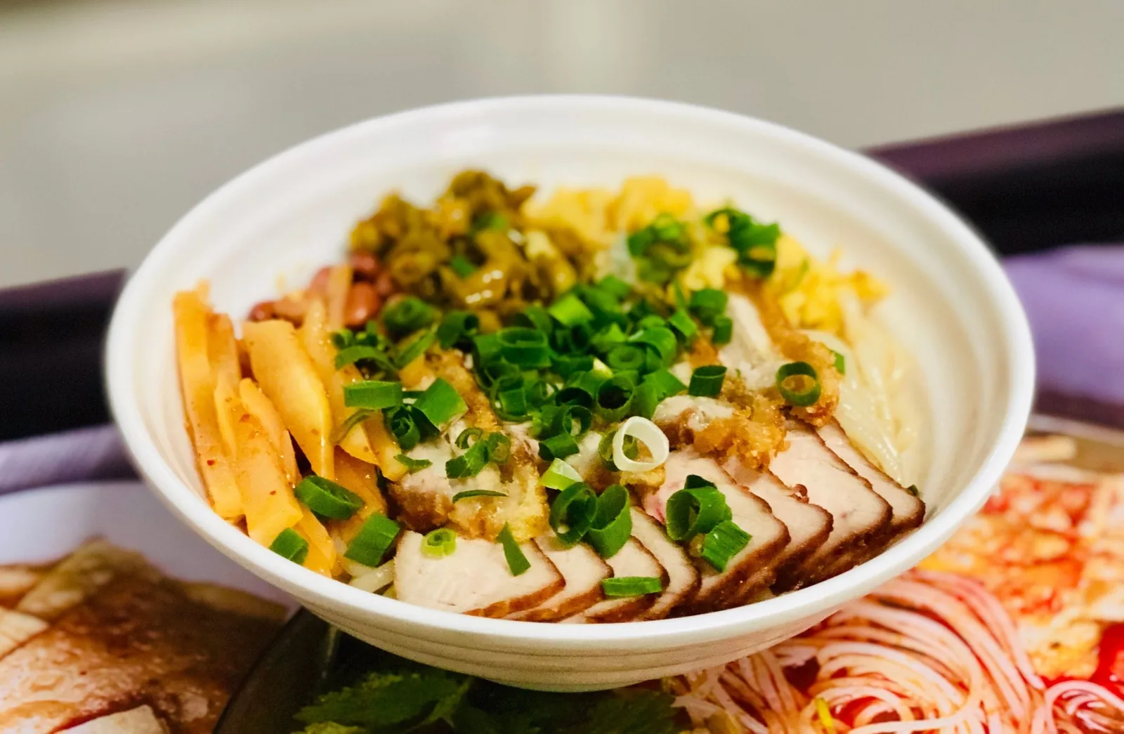 A white bowl of noodles topped with sliced pork, scallions, egg, and pickled vegetables, a specialty in Phu Quoc.