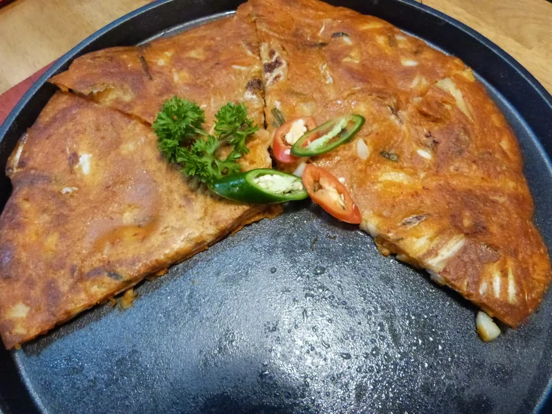 A sliced savory pancake with seafood and kimchi, garnished with chili and parsley, served on a dark plate in Ho Chi Minh.