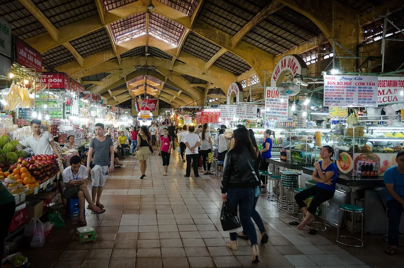 Shoppers walk through the bustling arched interior of Ben Thanh Market with food stalls and vendors in Ho Chi Minh City.