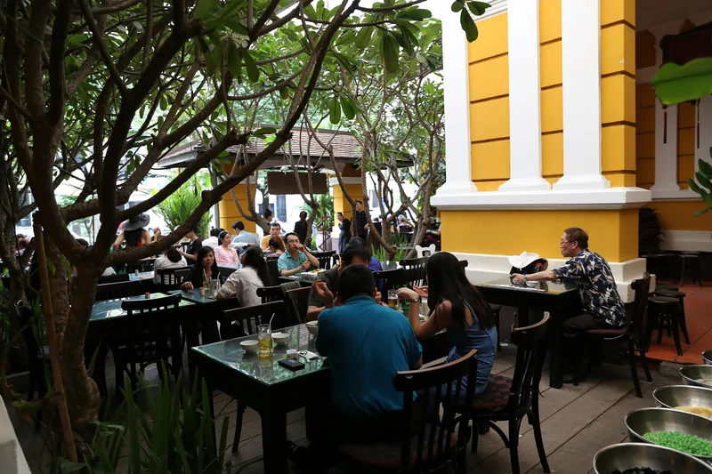 Diners relax at outdoor tables under frangipani trees beside a yellow colonial building in Ho Chi Minh City.